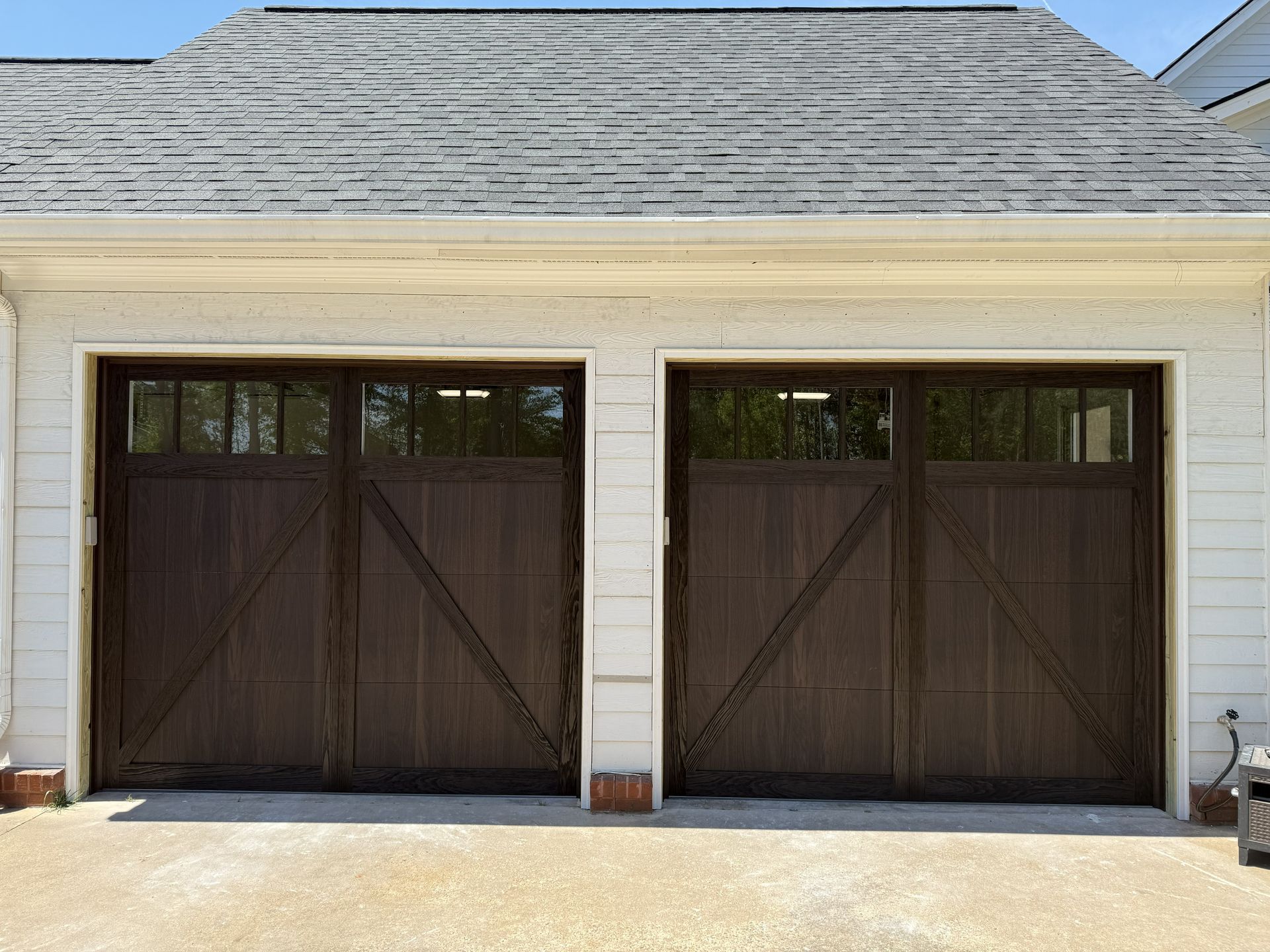 Two brown wooden garage doors with windows above, on a white building, gray shingle roof.