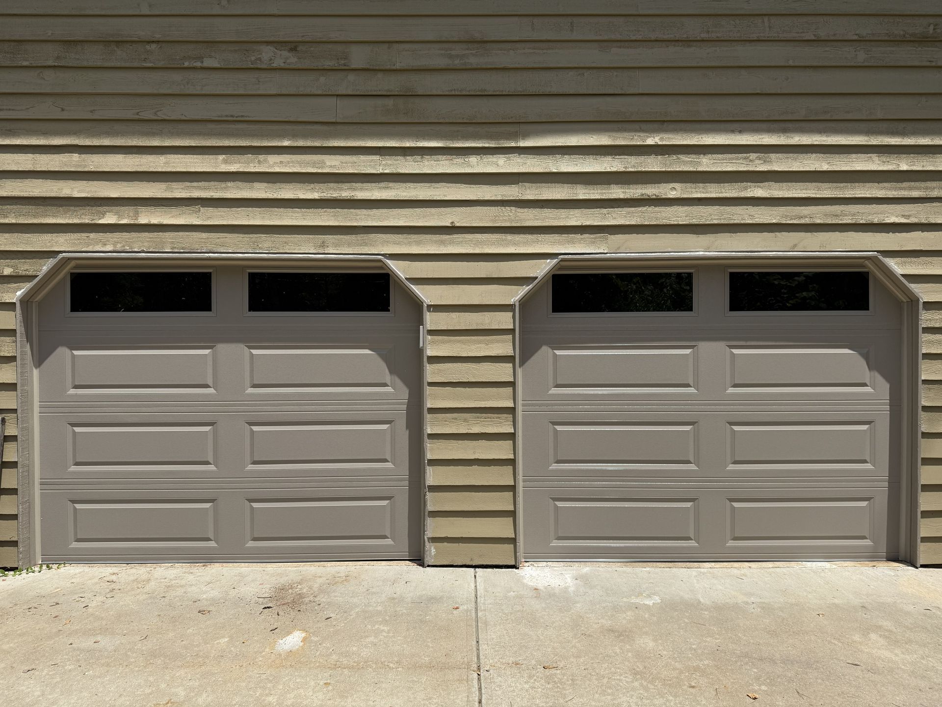 Two tan garage doors with black-paned windows, side-by-side, against a tan horizontal-plank wall.