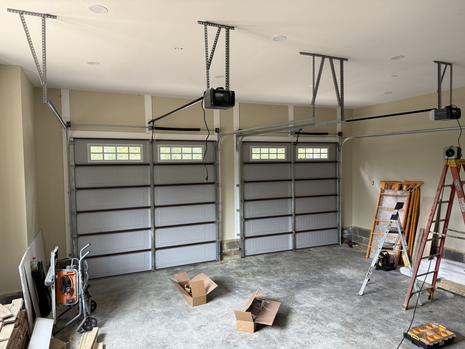 Garage interior with two gray garage doors, installed with automatic openers, boxes, and tools present.