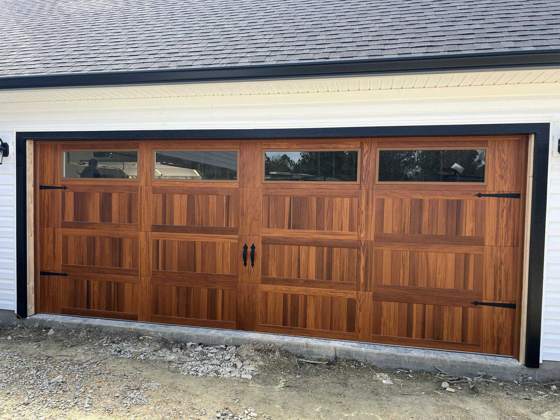 Wooden garage door with rectangular windows, black hardware, and wood-grain texture; beneath a white house.