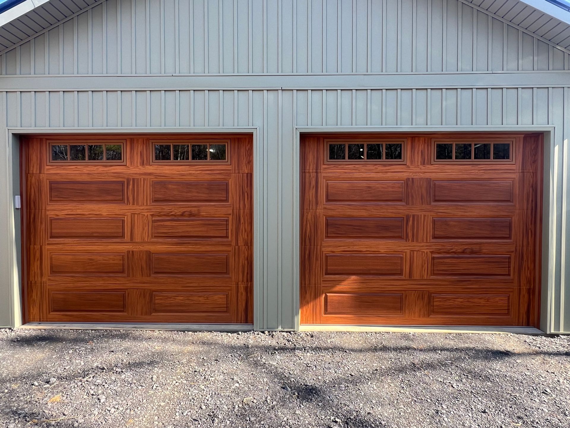 Two brown wooden garage doors with windows, set in a light green building. Gravel below.