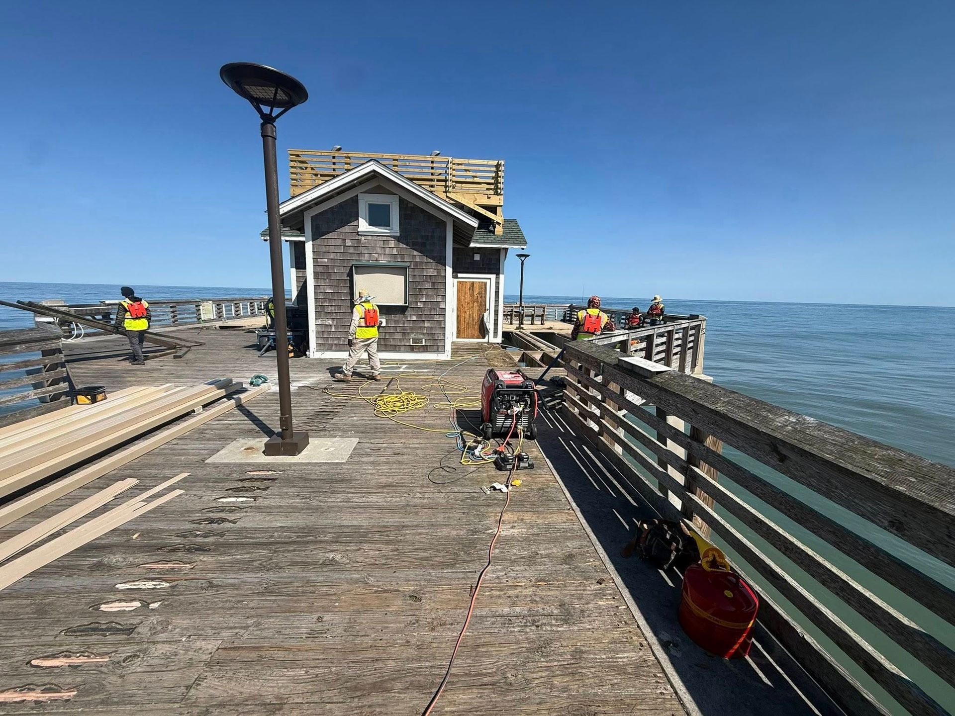 Sunlit pier with a small building, benches, fishing gear, and ocean on both sides.