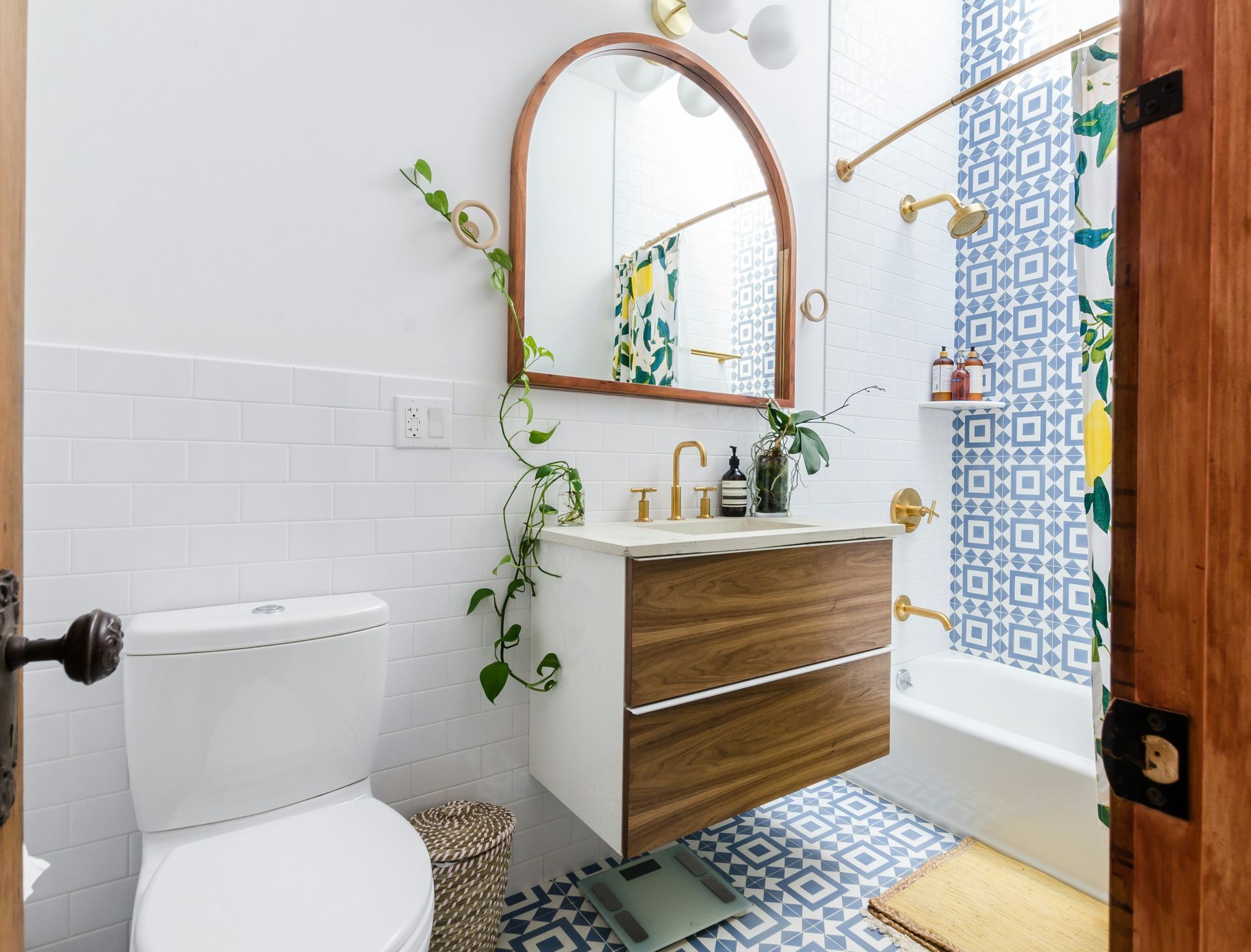 Bright bathroom with white toilet, wood vanity, arched mirror, and blue patterned shower curtain and floor tile
