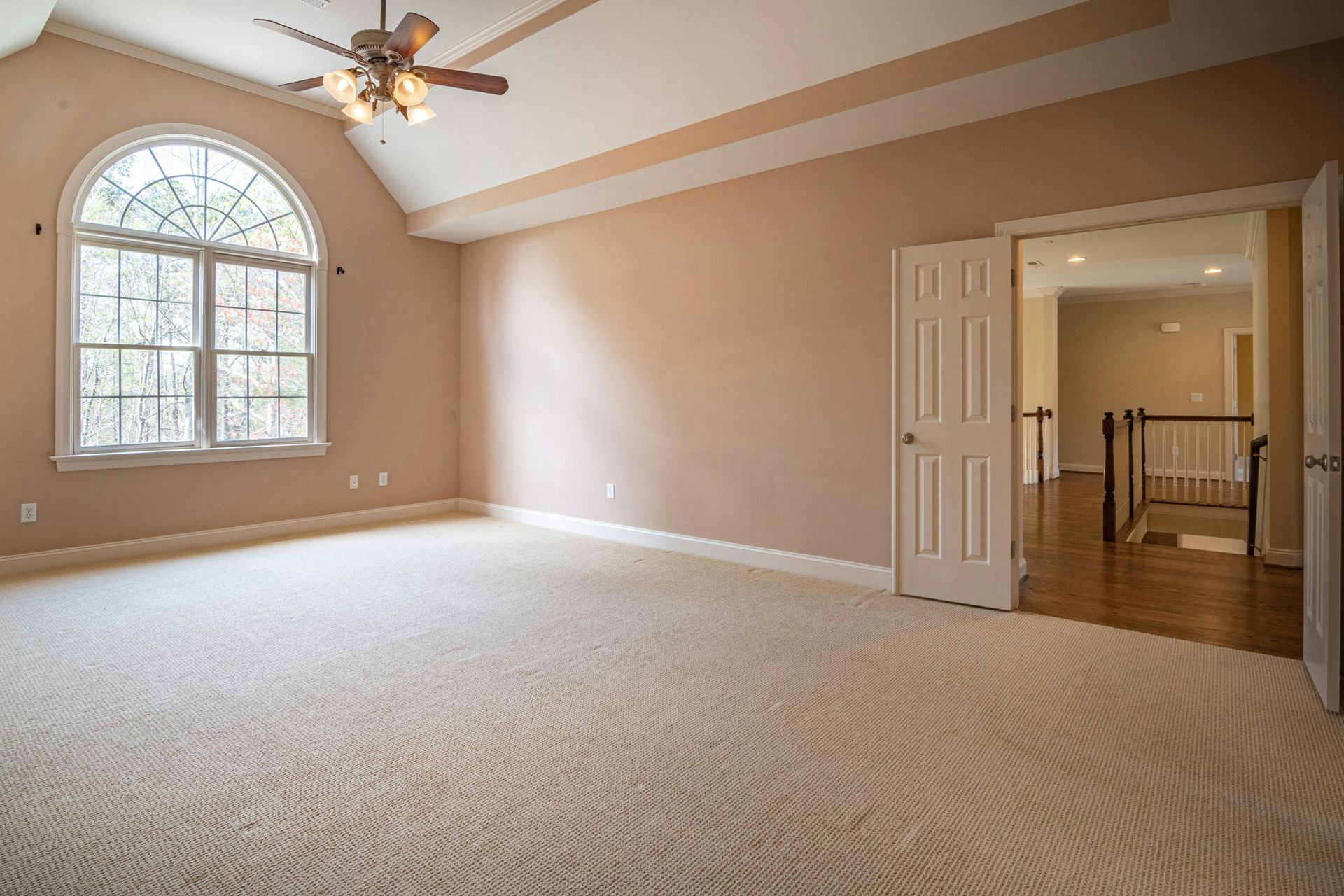 Spacious beige carpeted bedroom with arched window, ceiling fan, and open double doors to a hallway