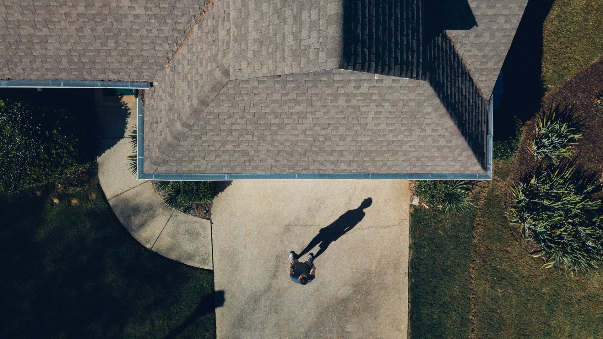 Aerial view of a person walking across a driveway beside a house with a shingled roof.