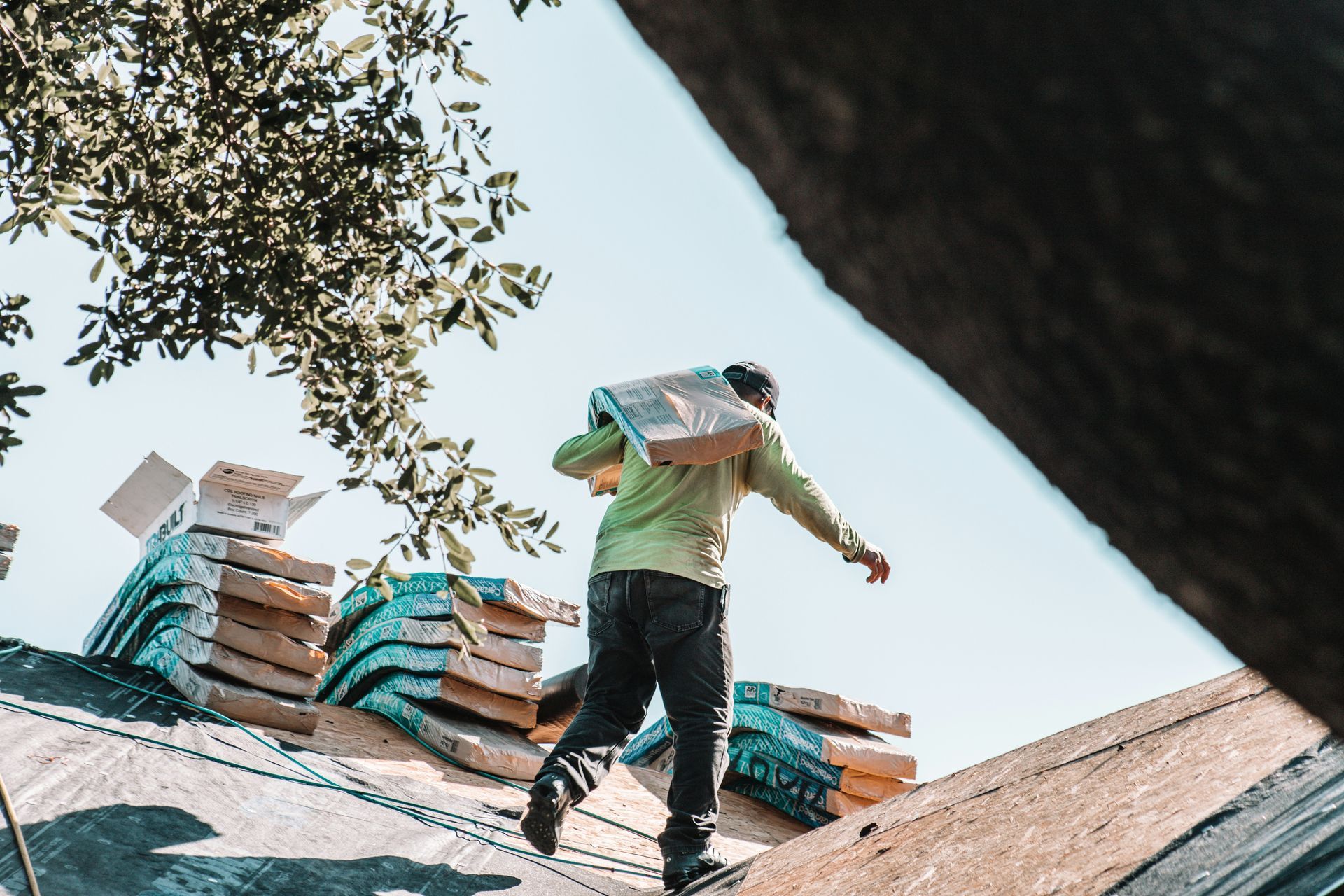 Worker carrying roofing materials on a sloped roof under a tree