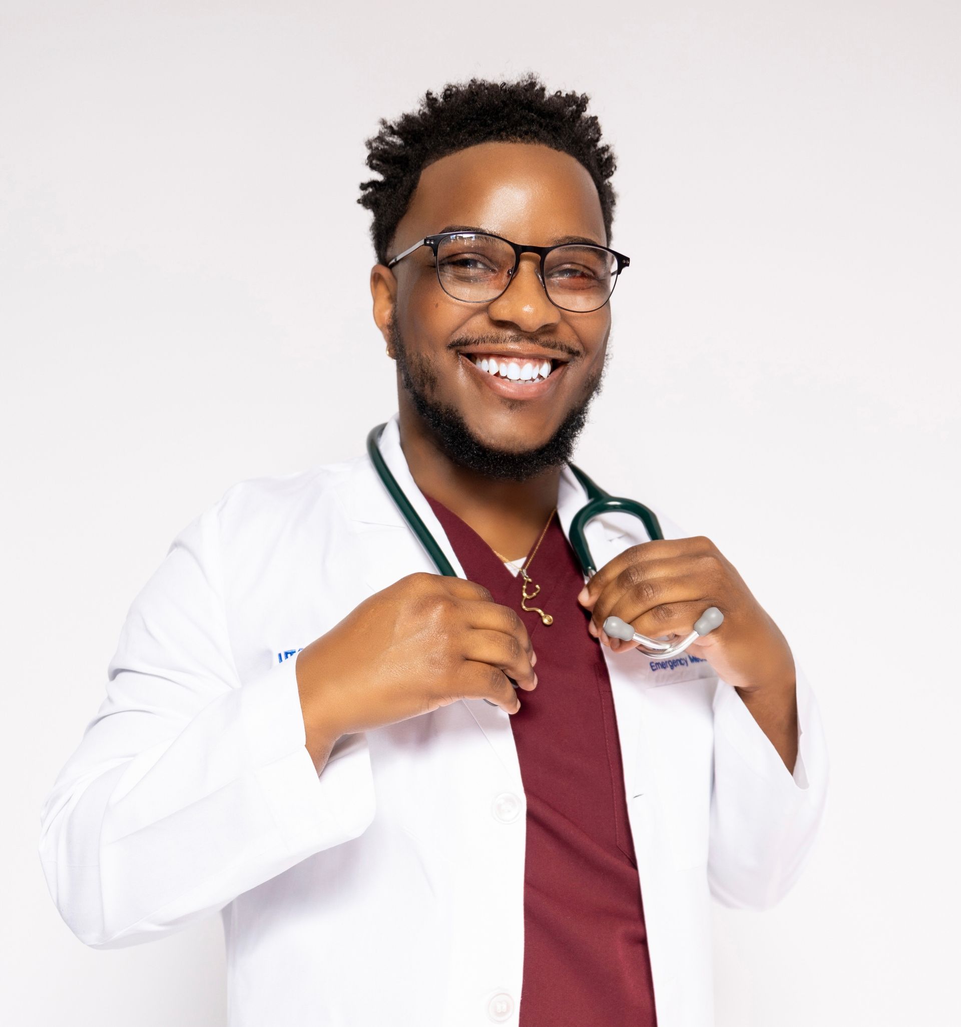 Smiling person in medical attire, stethoscope, and glasses. Burgundy scrubs, white coat, holding coat.