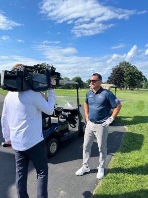 A man is standing next to a golf cart on a golf course talking to a cameraman.