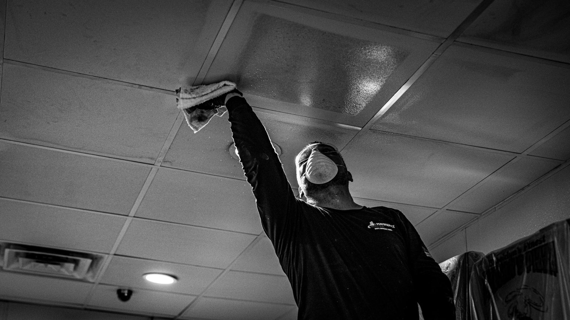 A man wearing a mask is cleaning the ceiling with a cloth.