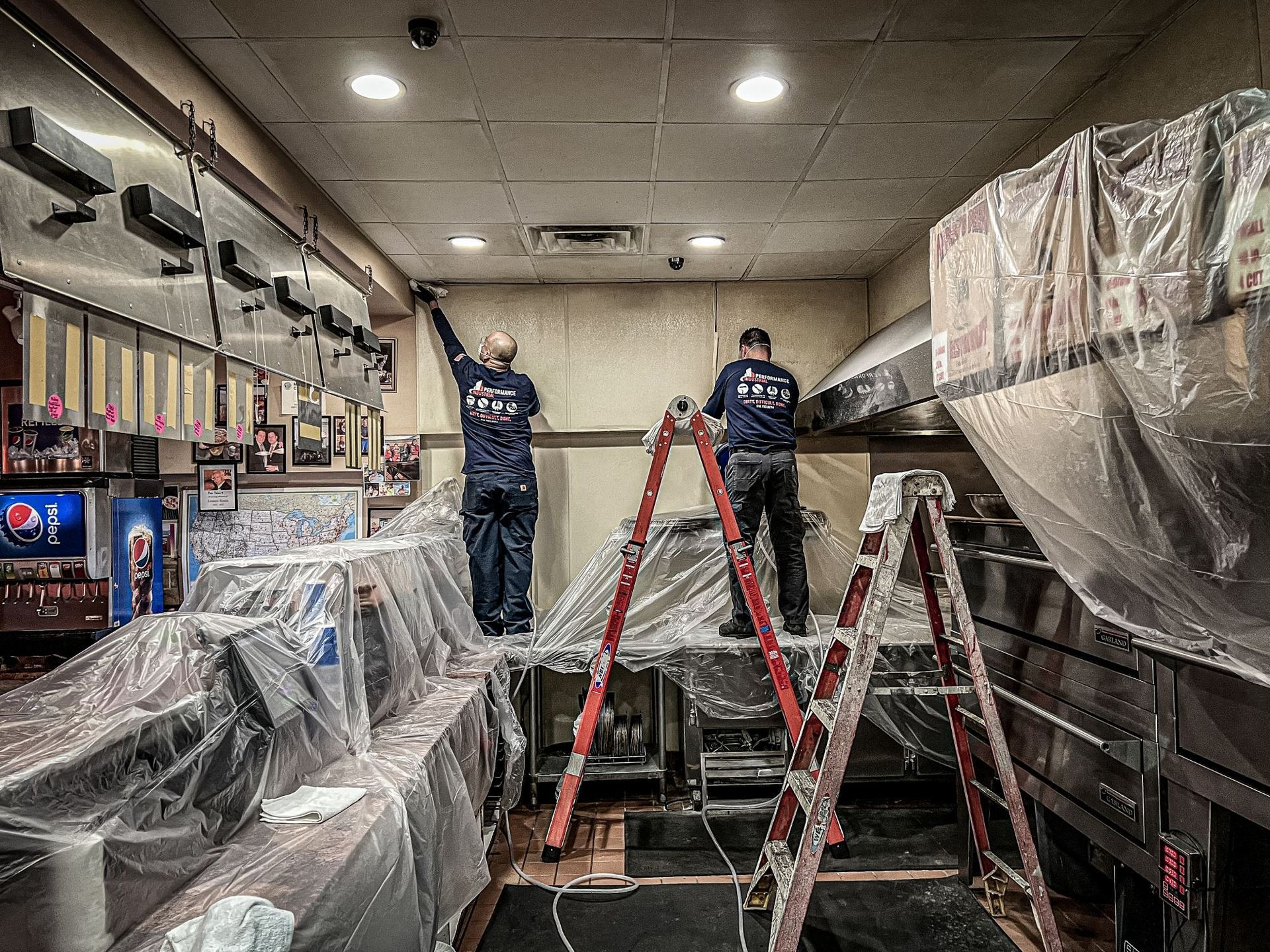 Two men are standing on ladders in a kitchen covered in plastic.