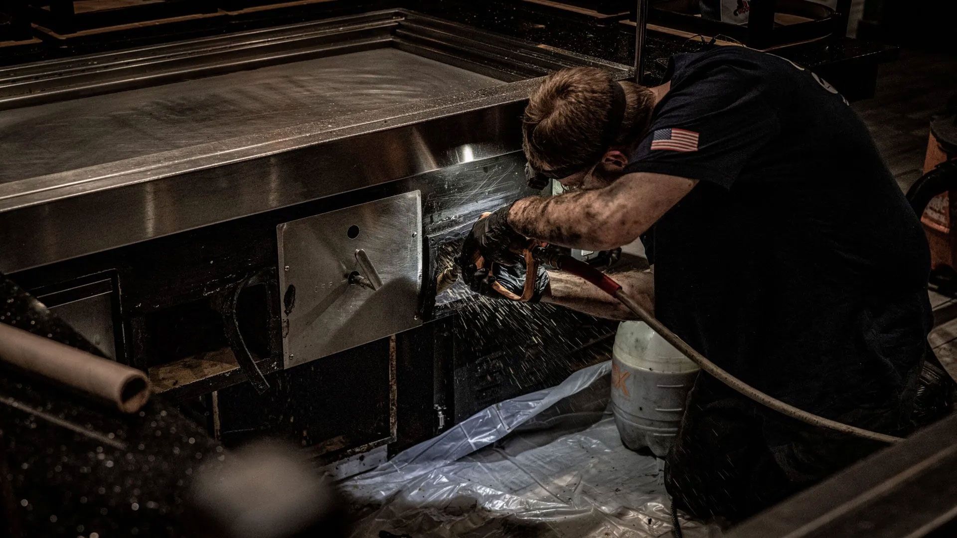 Worker using power tool to cut metal near a large stainless steel surface.