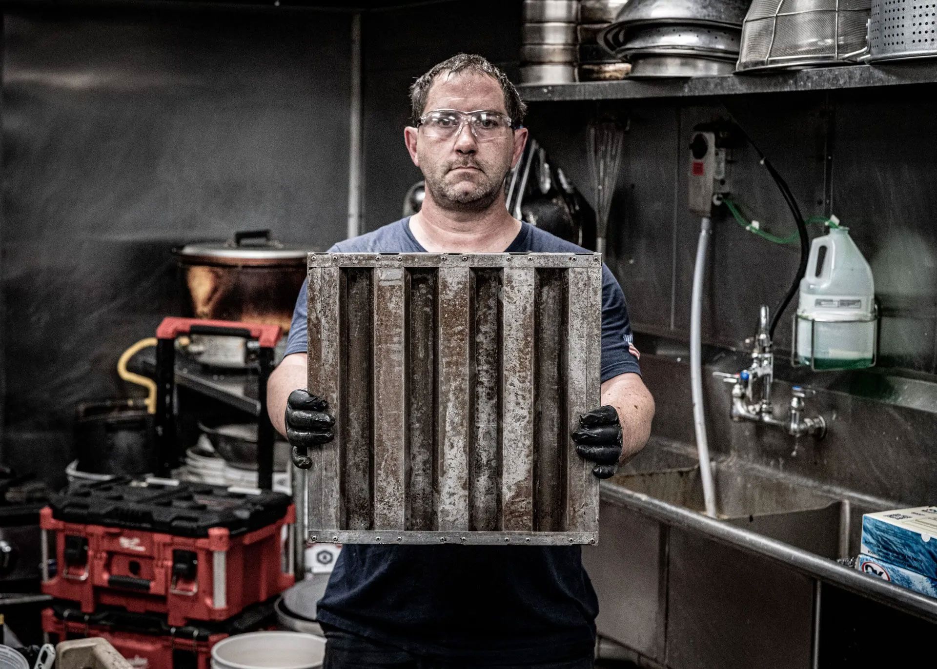 A man holds a grid from a kitchen exhaust, inside a cleaning station.