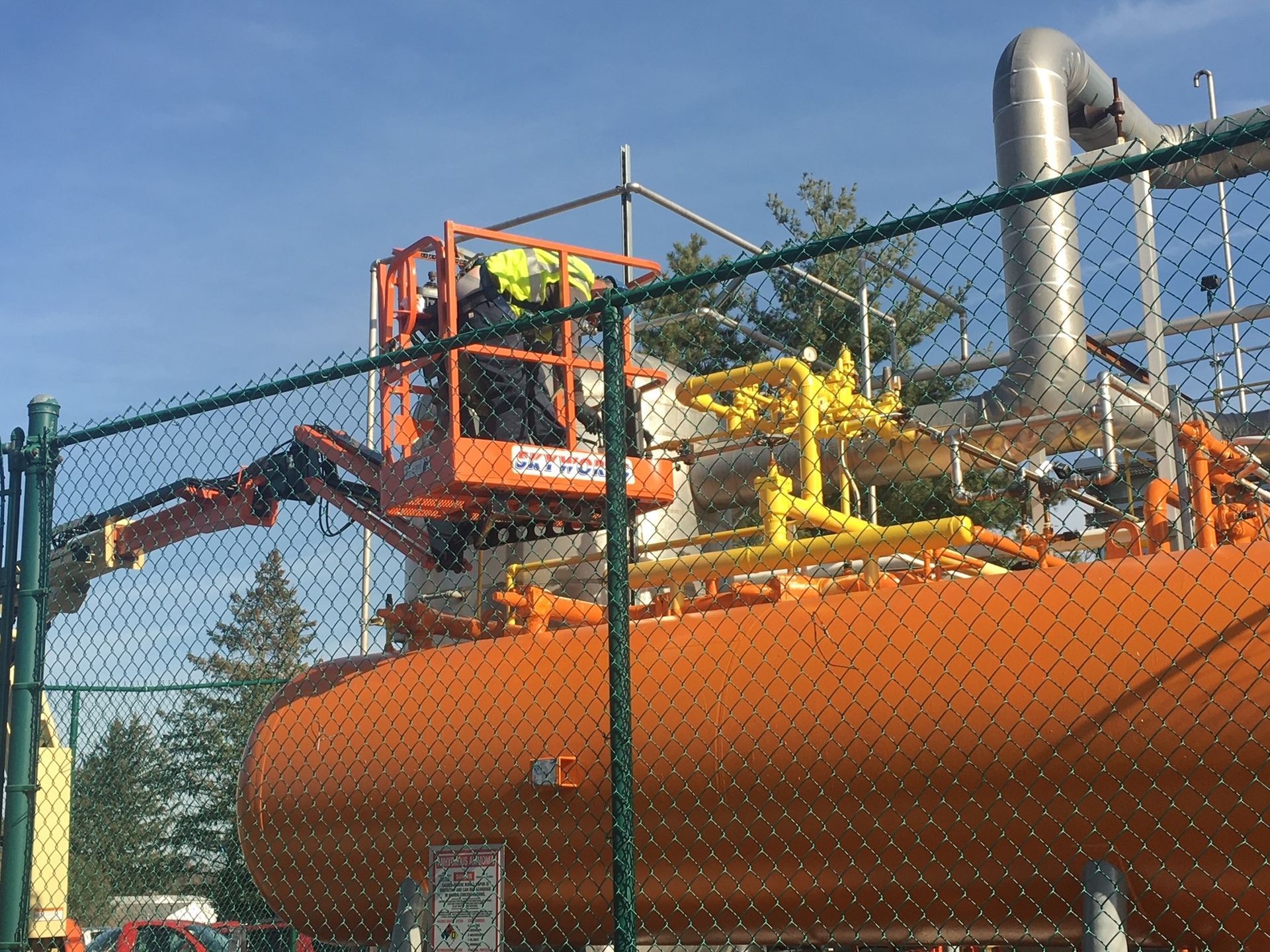 A man in a crane is working on a large orange tank behind a chain link fence.