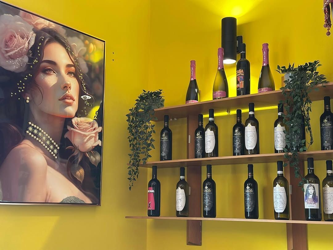 Una parete colorata espone bottiglie di vino su scaffali di legno, accanto al ritratto floreale di una donna.