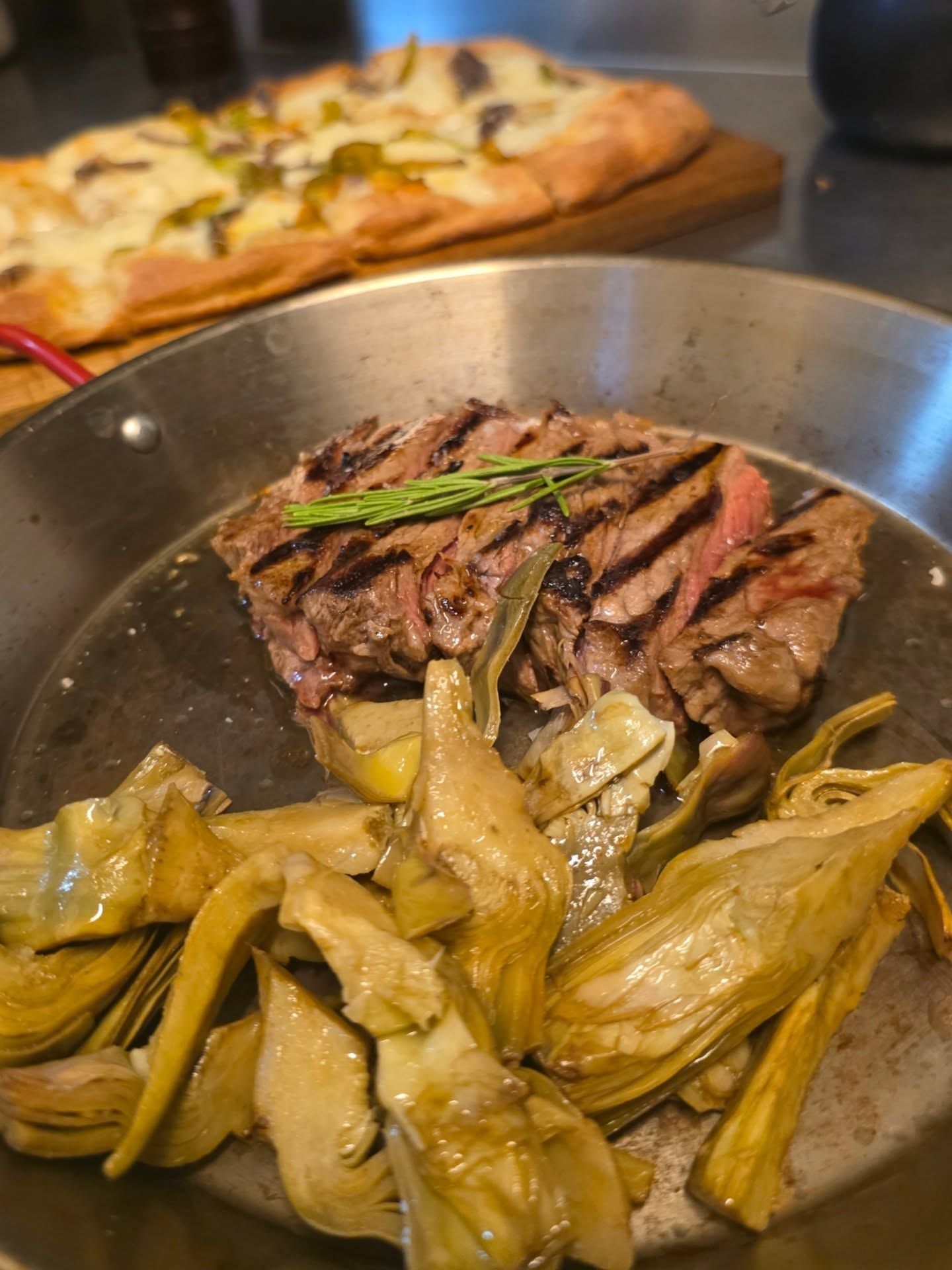 Bistecca alla griglia con carciofi in padella, pizza sullo sfondo.