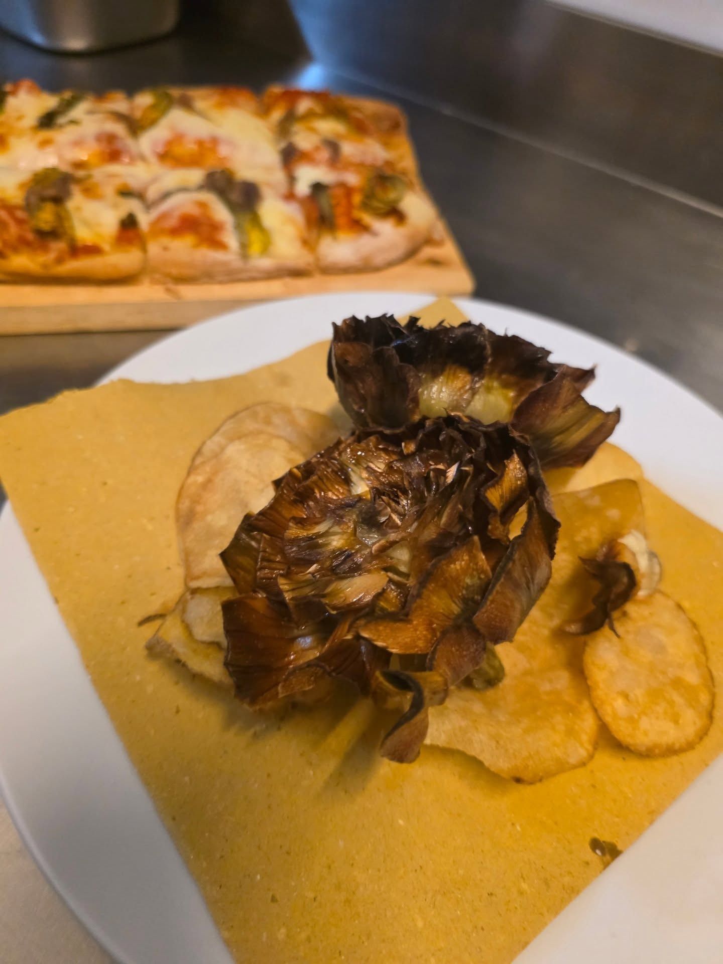 Carciofi fritti e patatine fritte su un foglio di carta gialla, con una pizza rettangolare sullo sfondo.