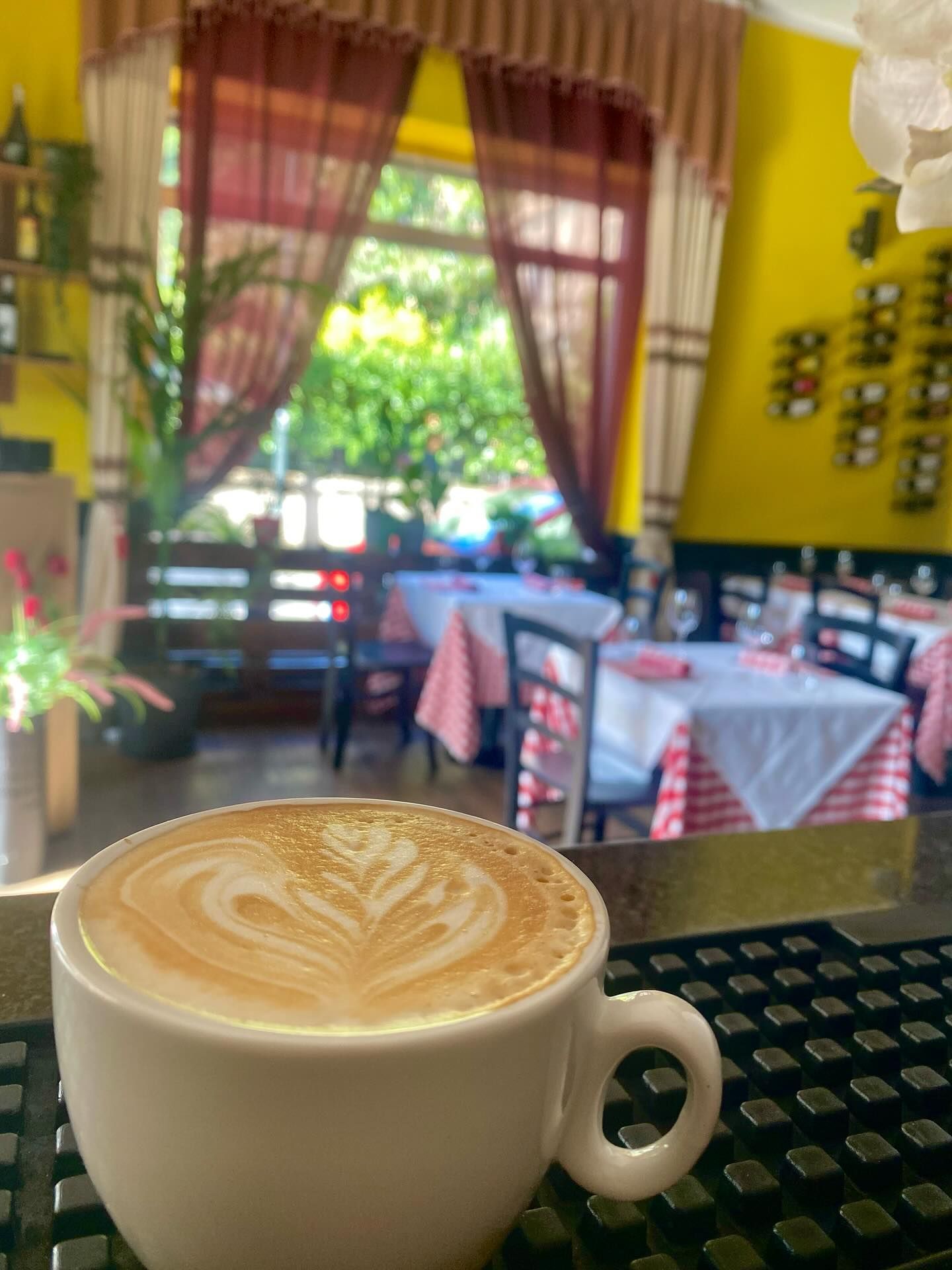 Latte macchiato con latte art sul bancone di un bar, sullo sfondo l'interno di un ristorante con tavoli e tovaglie a quadri rossi.