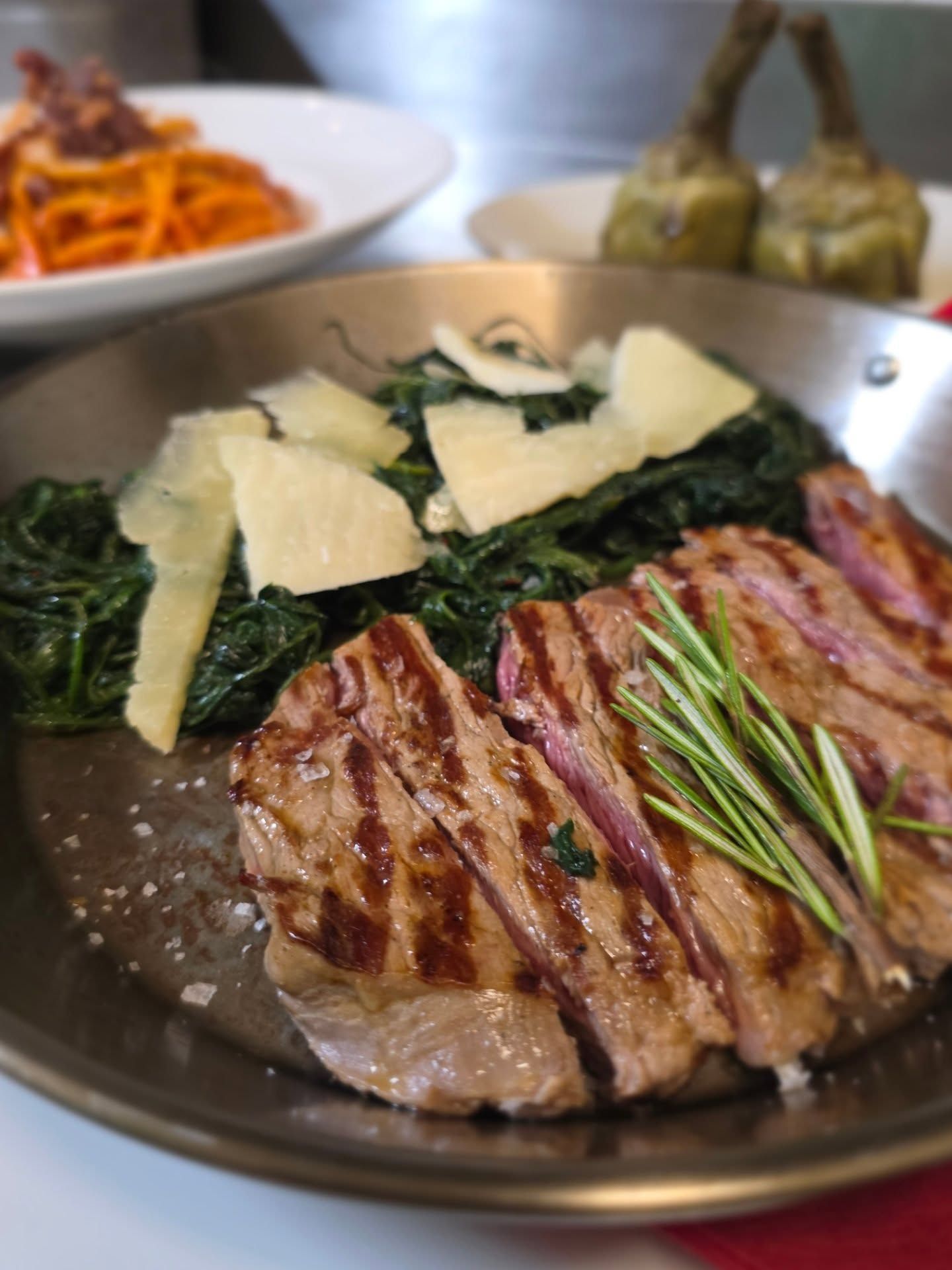 Bistecca alla griglia con spinaci e parmigiano, guarnizione di rosmarino, in una padella di metallo. Pasta e carciofi sullo sfondo.