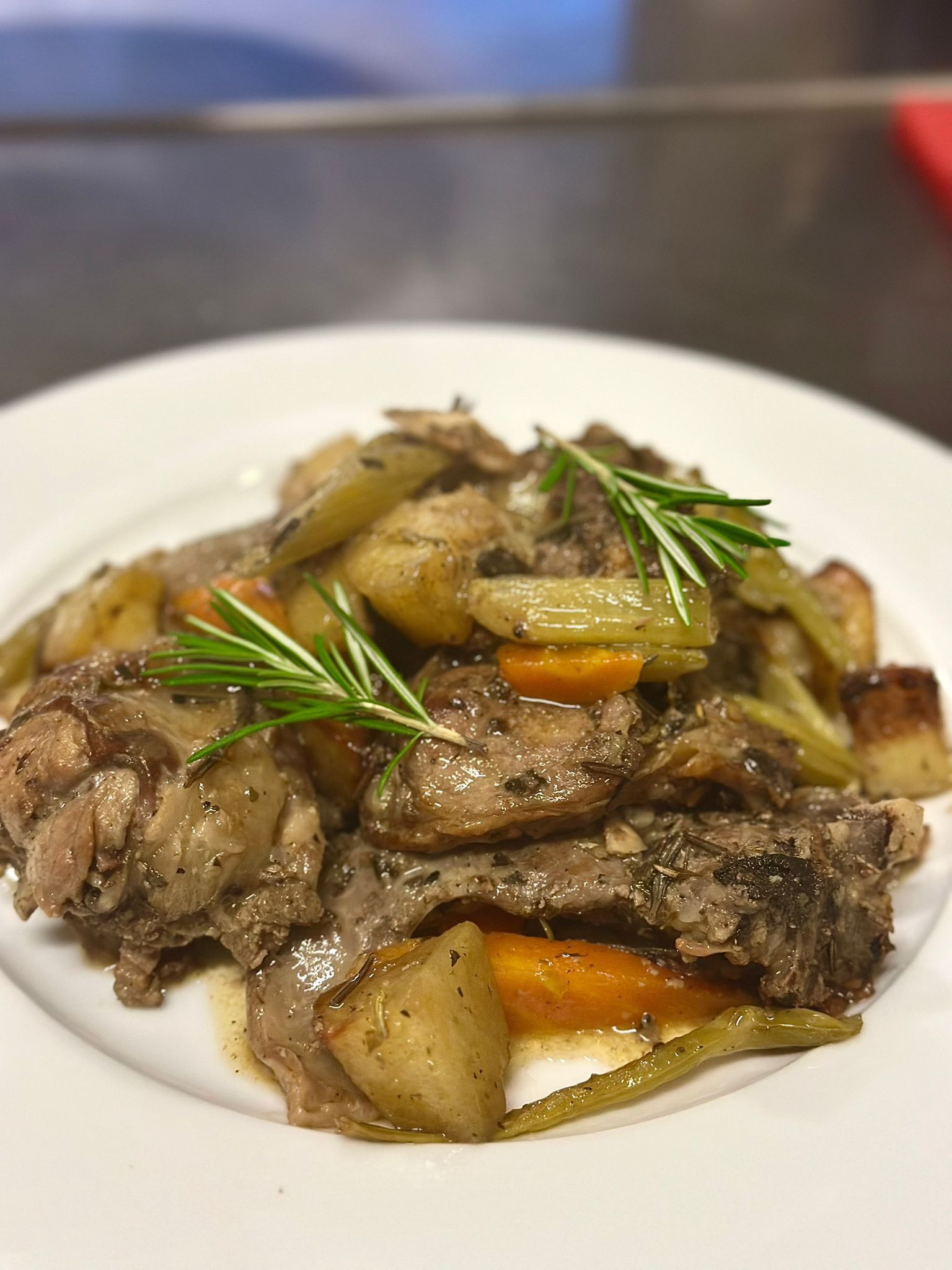 Carne stufata con patate, carote e sedano su un piatto bianco, guarnita con rosmarino.