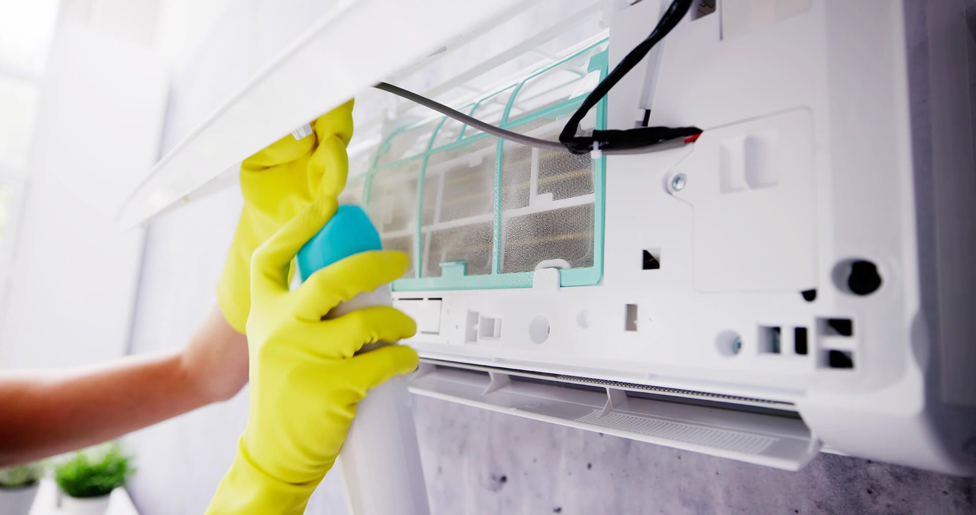 Person in yellow gloves cleaning an air conditioner filter with a spray bottle.