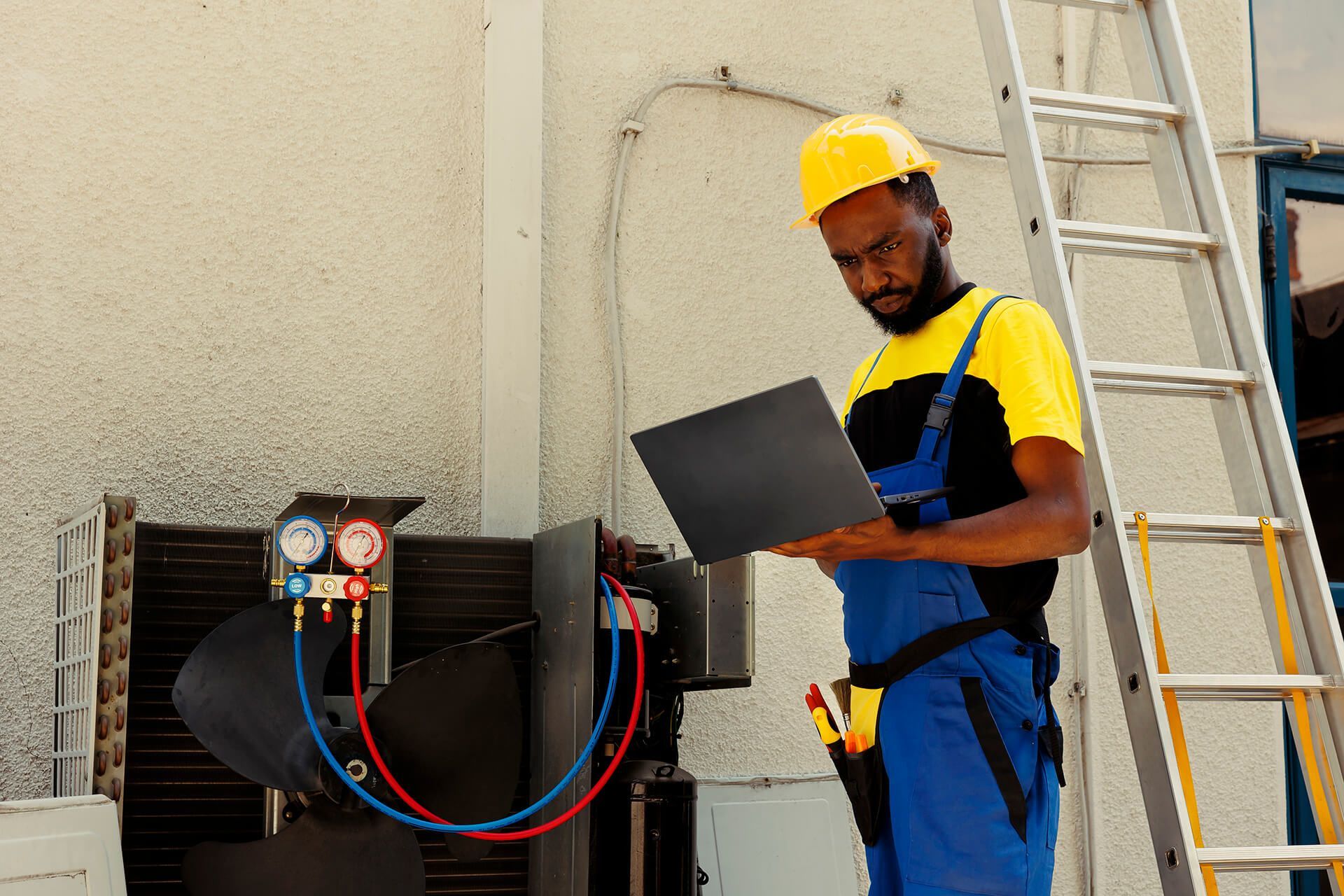 HVAC technician in yellow hard hat and blue overalls using a laptop to inspect machinery on a building rooftop.