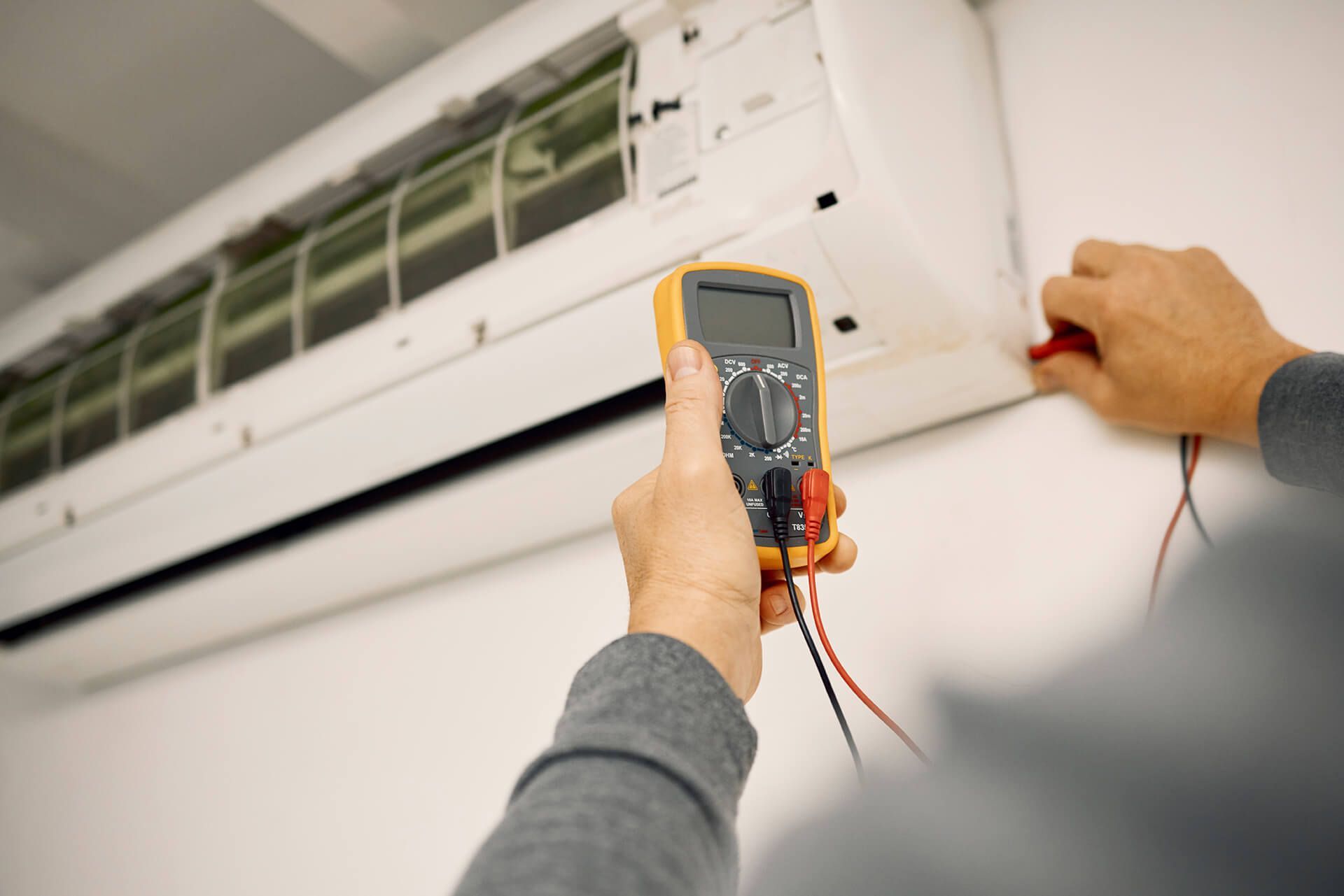 Person using a multimeter to inspect an air conditioning unit; hands hold the device and probes.