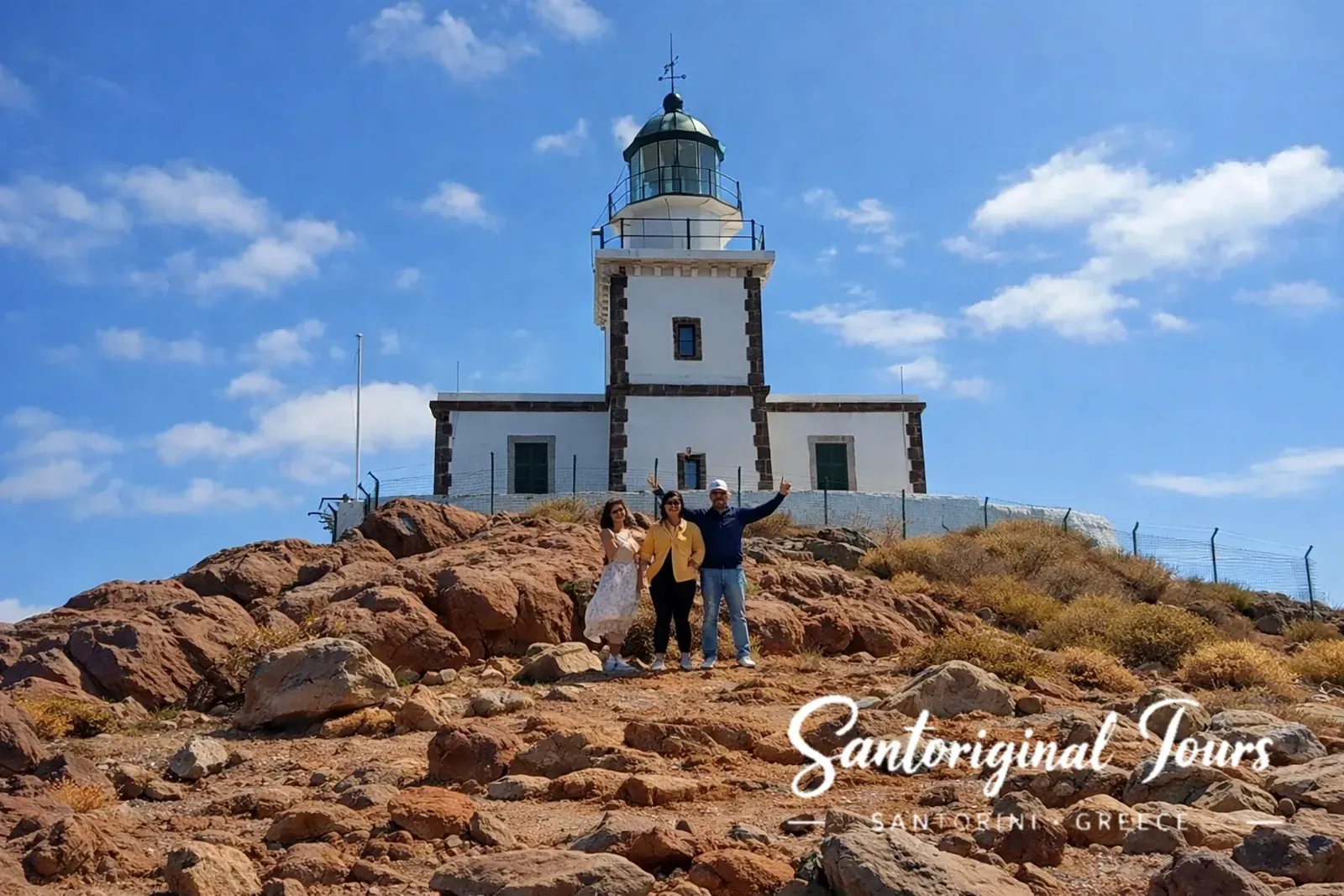 Our guests standing on the rocky hillside below Akrotiri Lighthouse in Santorini under a blue sky with scattered clouds.