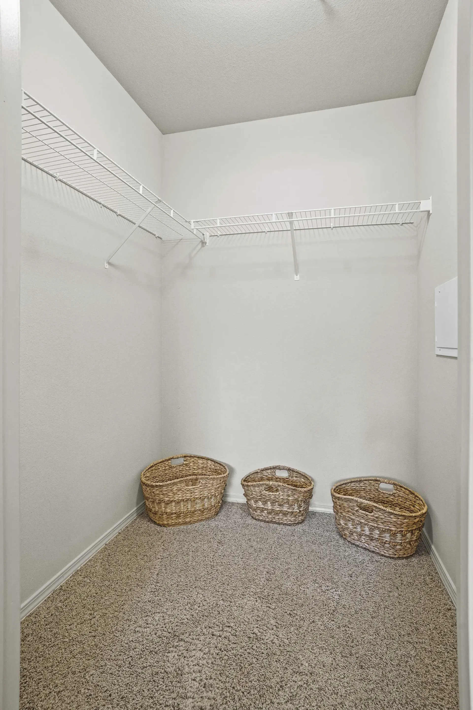 Bright walk-in closet with white wire shelving on two walls and three wicker baskets on beige carpet.