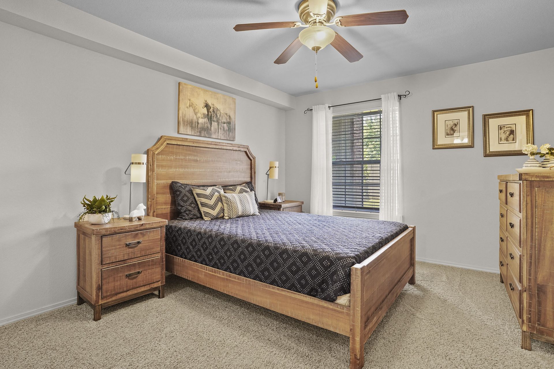Bedroom in an apartment with a wooden bed, two nightstands, dresser, ceiling fan, and a window with white curtains.
