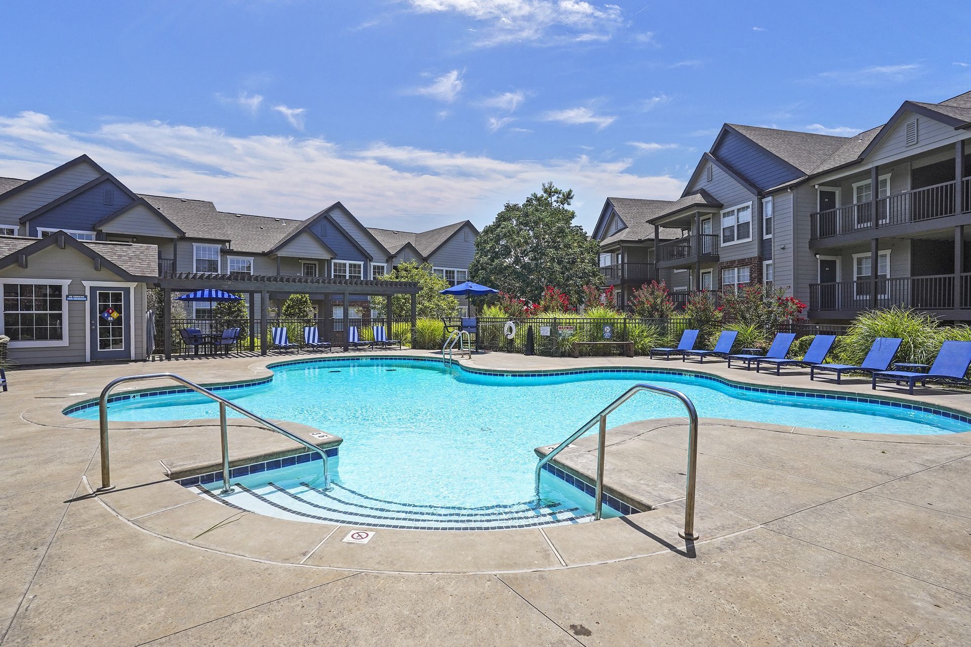Swimming pool in front of apartment buildings on a sunny day with blue sky and lounge chairs.