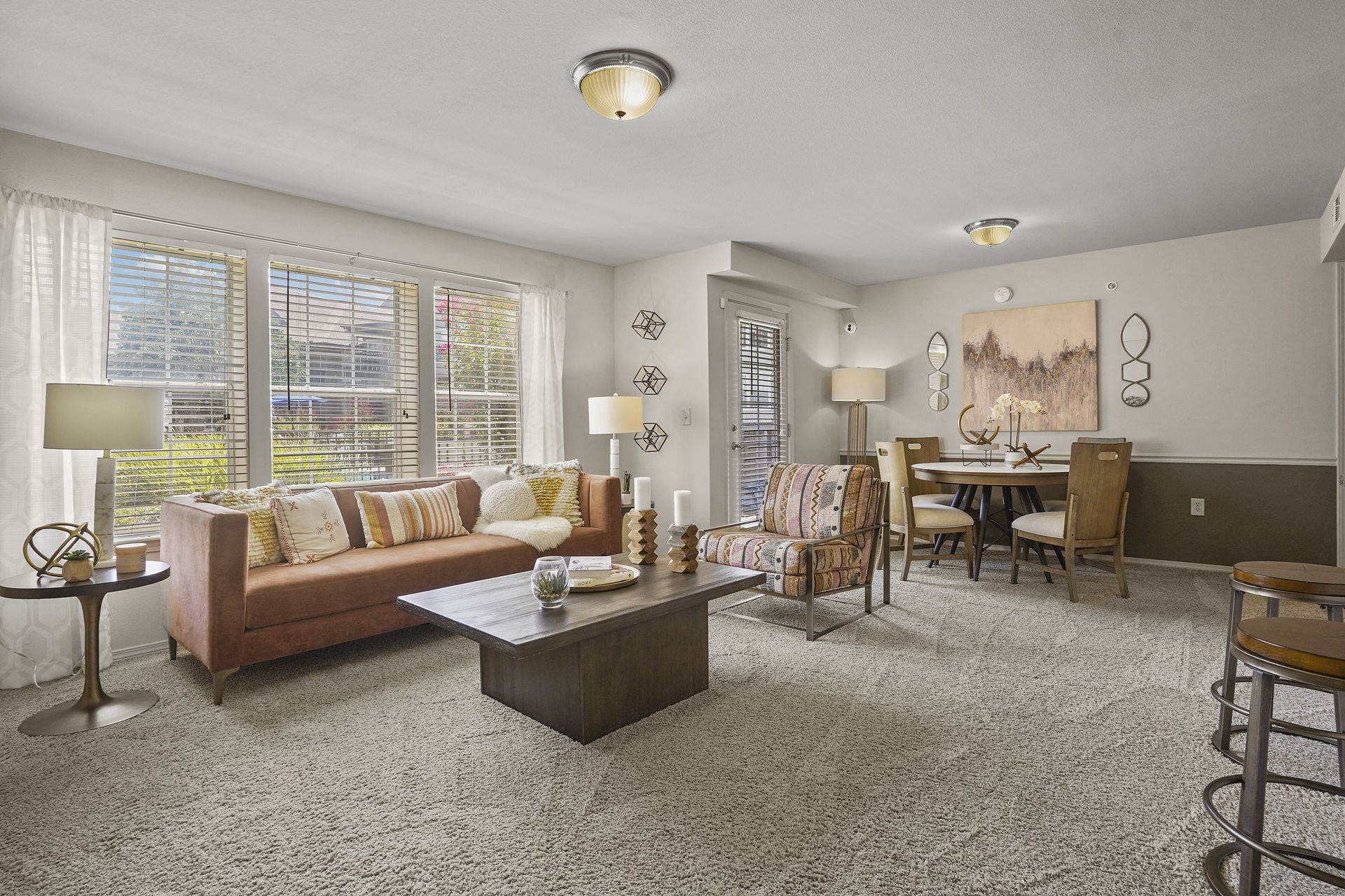 Living room with sofa, coffee table, dining table, and bar stools. Large window, patterned carpet.