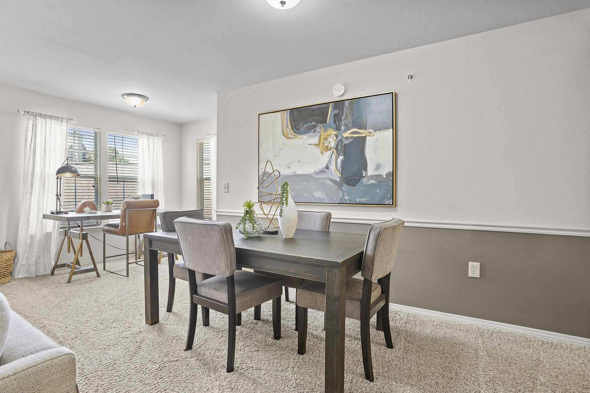 Interior view of a sunlit apartment showing a dining table with chairs and a desk near windows.