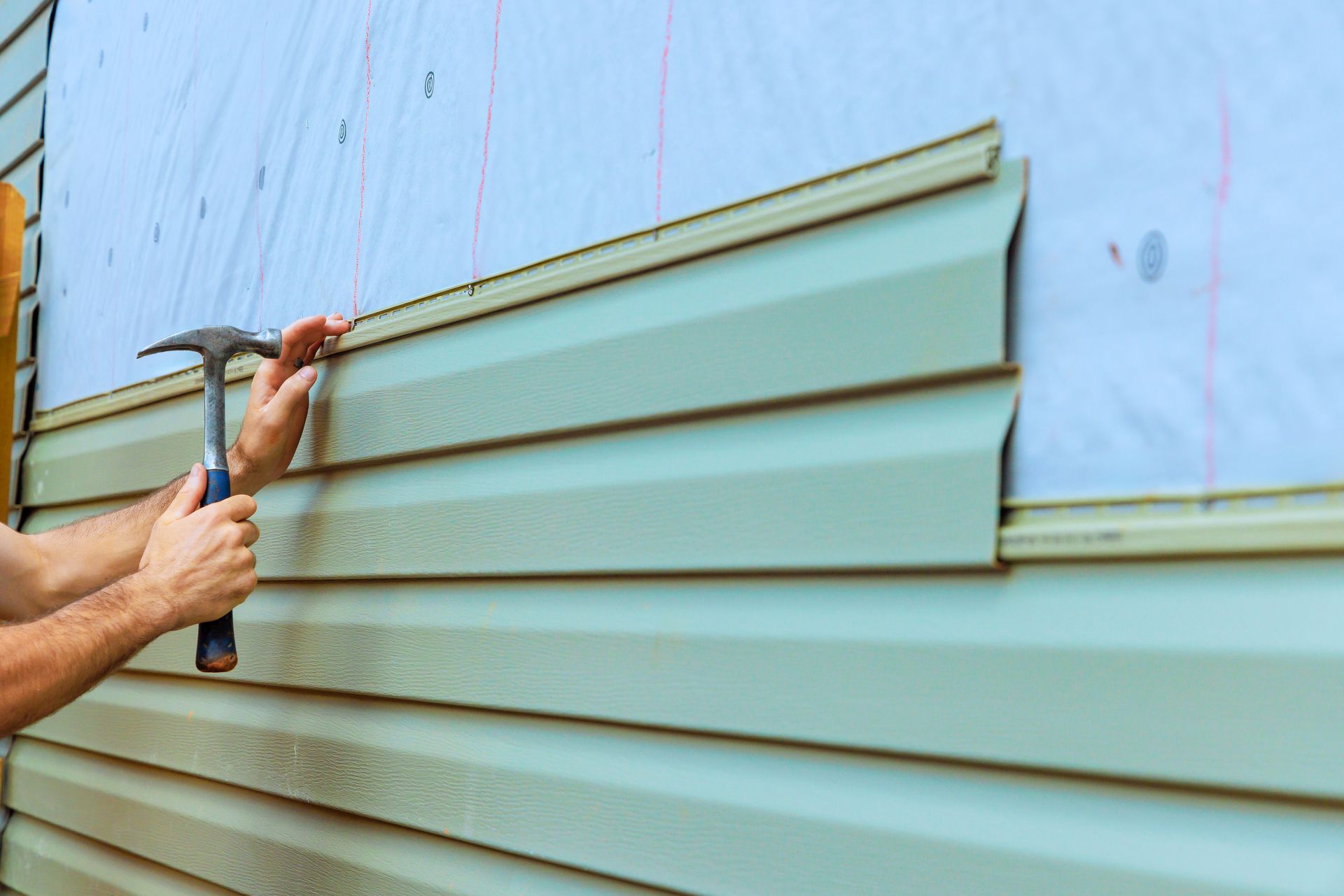 Hands installing pale green siding panels on an exterior wall with a hammer and pry bar