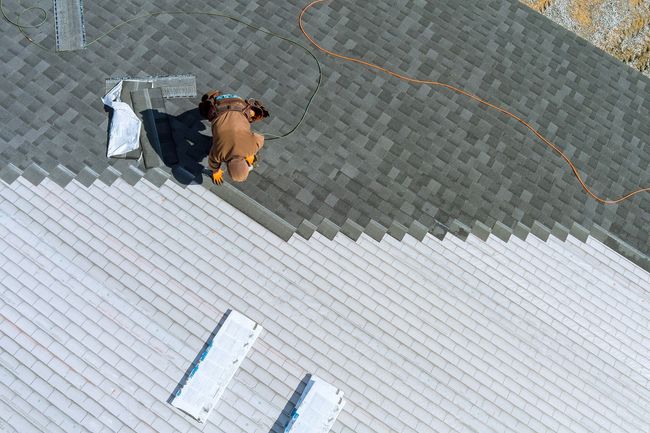 Worker kneeling on gray roof next to orange rope and tools near white roof edge