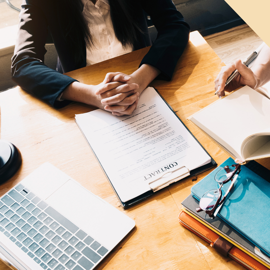 People at a table with a contract, laptop, and notebook. One person clasps hands, the other writes.