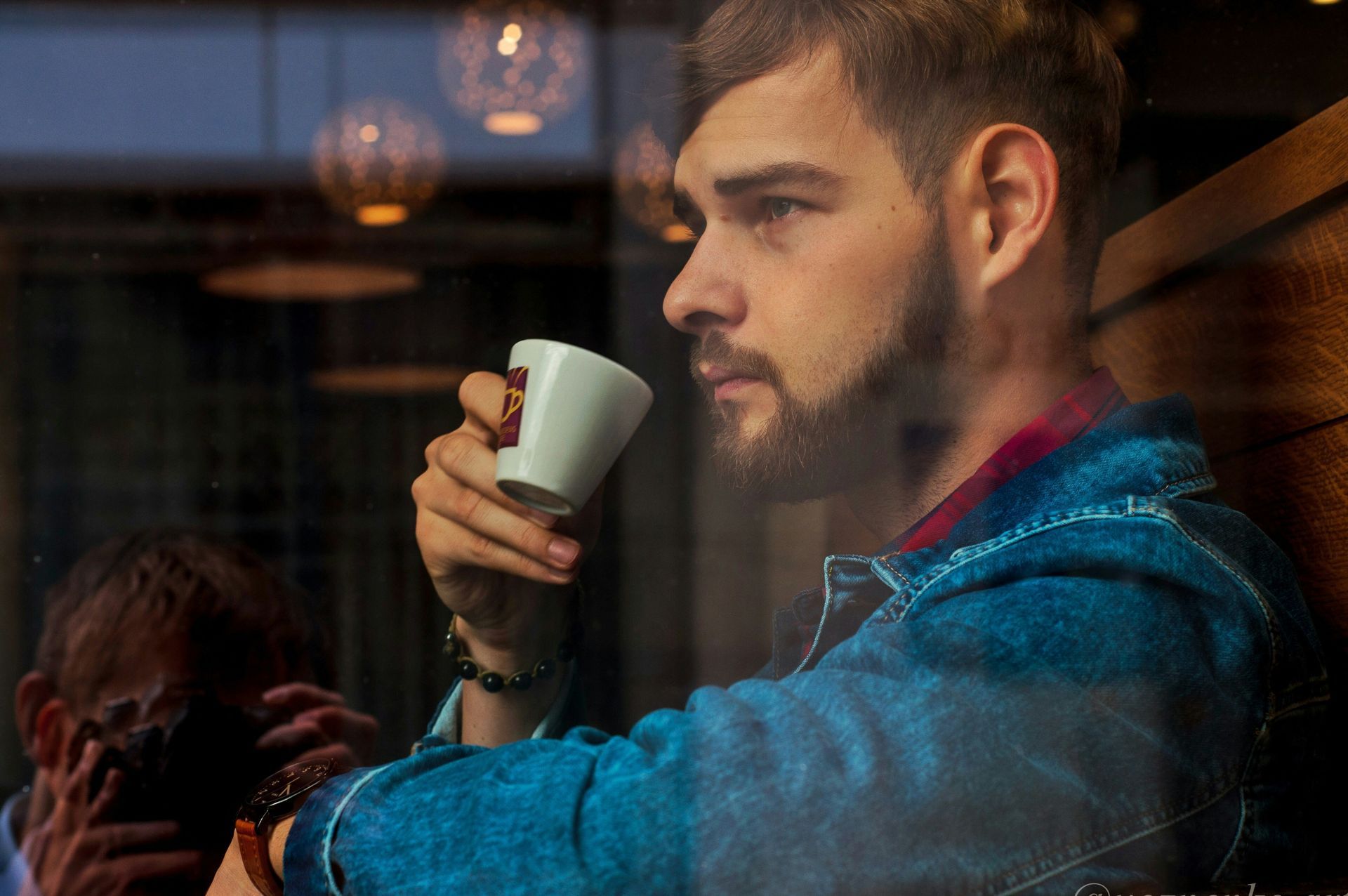 Un hombre con una chaqueta vaquera azul bebe de una taza blanca, mirando por una ventana; entorno de cafetería.