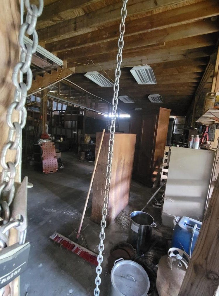 Interior of a cluttered workshop with wooden structures, metal chains hanging, various containers, and overhead fluorescent lights.
