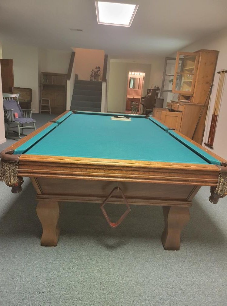 Pool table with green felt in a room. Wooden furniture, stairs, and a light fixture are visible.