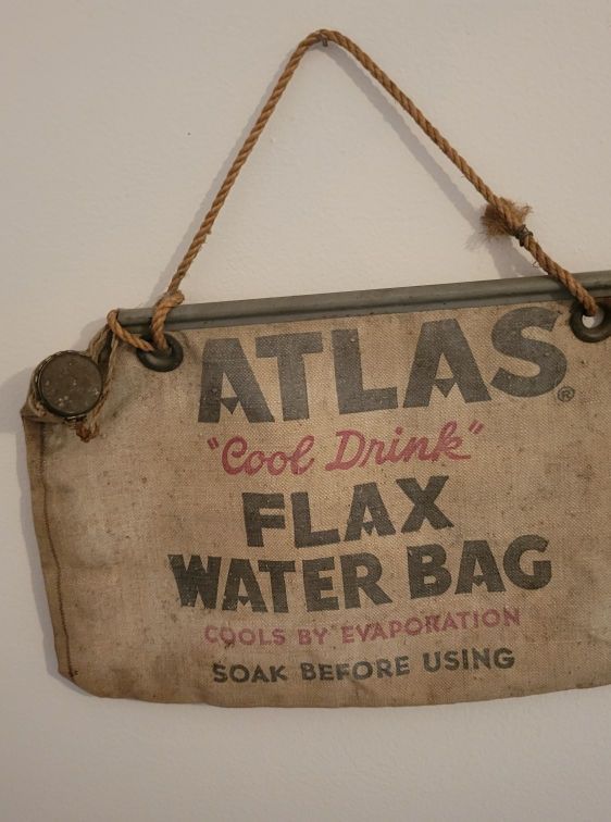 Tan Atlas flax water bag hanging on a white wall, with rope handle.