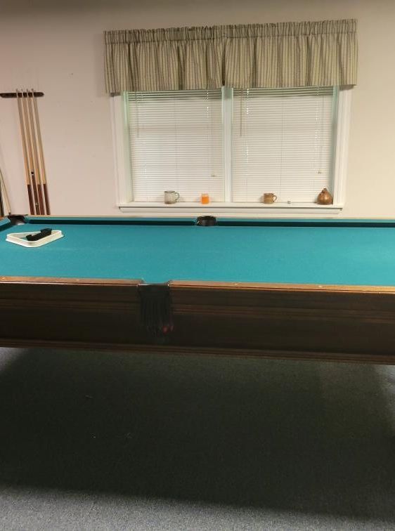 Pool table with green felt, window behind, cue sticks on the side, in a room.