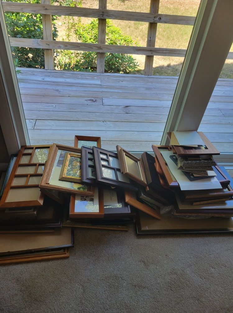 Pile of wooden picture frames of various sizes on a carpeted floor, window and deck in the background.