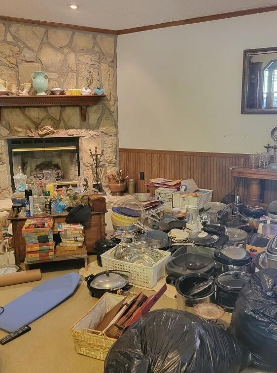 Room filled with items near a stone fireplace and light-colored walls. Piles of items and black trash bags on the floor.