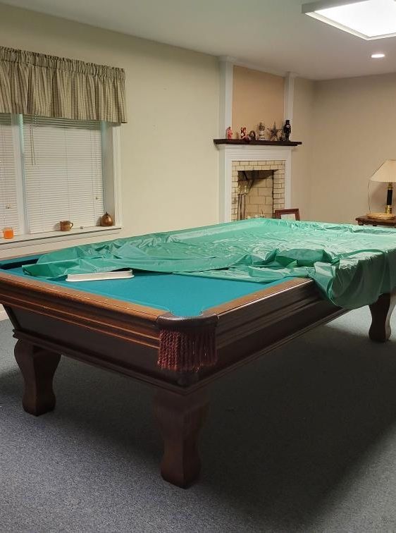 Pool table covered in a green tarp in a room with a window, fireplace, and lamp.