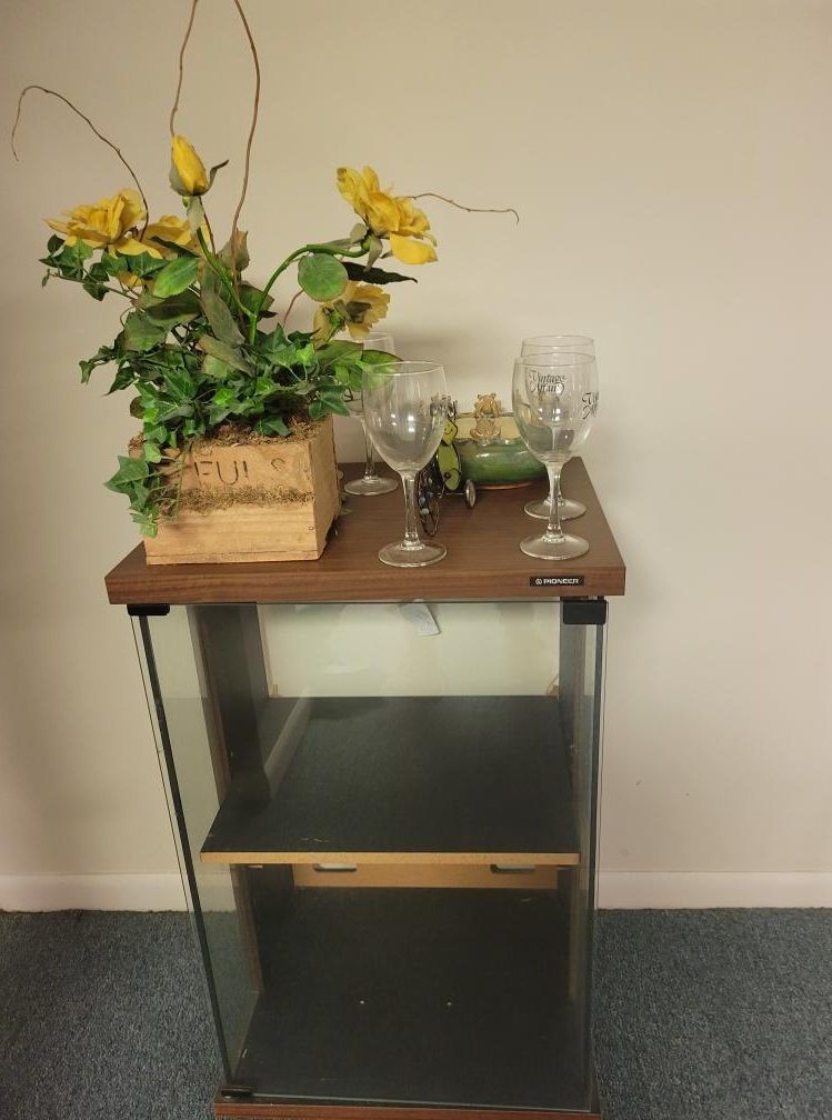 A wood and glass side table holds flowers, wine glasses, and a small dish against a white wall.