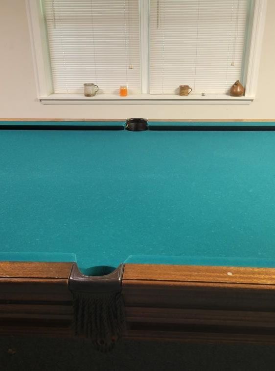 Pool table with teal felt, brown wooden rails, and a window with blinds in the background.