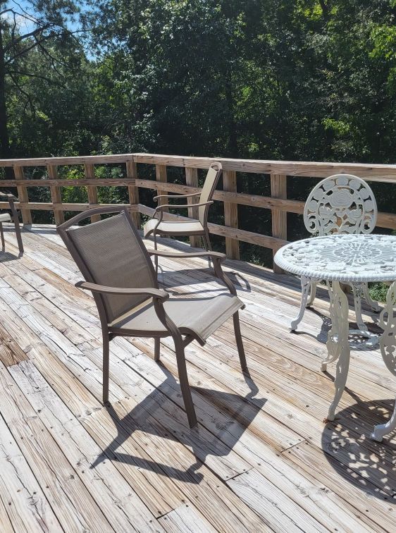 A sunny outdoor deck with wooden railing and various chairs, including a metal table with a white, patterned top.
