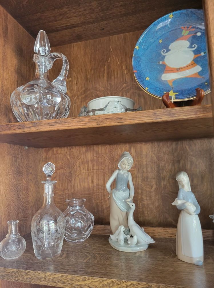 Wooden cabinet shelves with crystal decanters and figurines, including a girl with geese and a Santa plate.