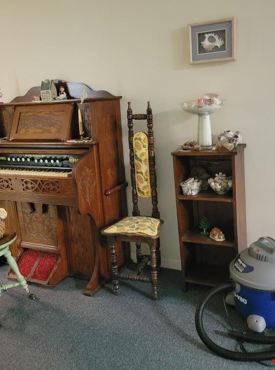 Antique organ, high-back chair, small bookcase with seashells, and a vacuum cleaner against a wall.