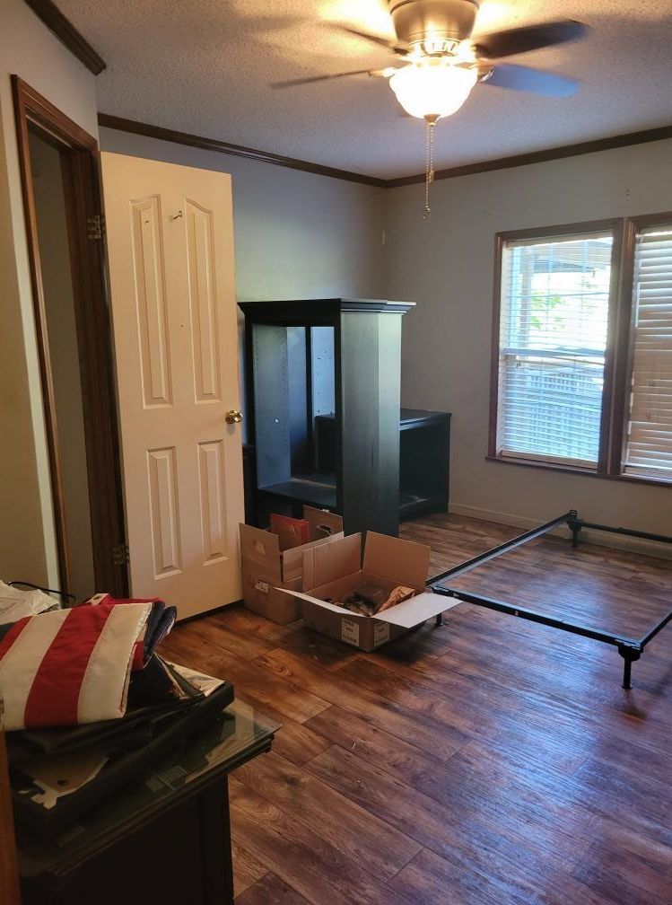 Bedroom with dark wood-look floor, bed frame, boxes, dark cabinet, white door, window with blinds.