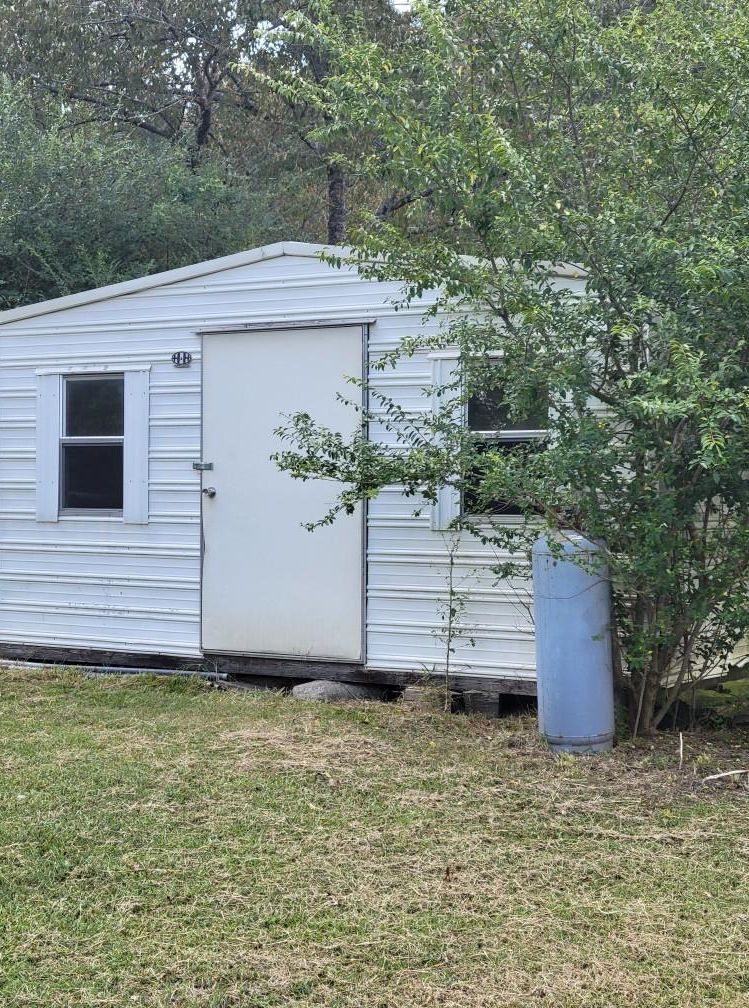White shed with closed door, small window, and overgrown foliage on a grassy lawn.